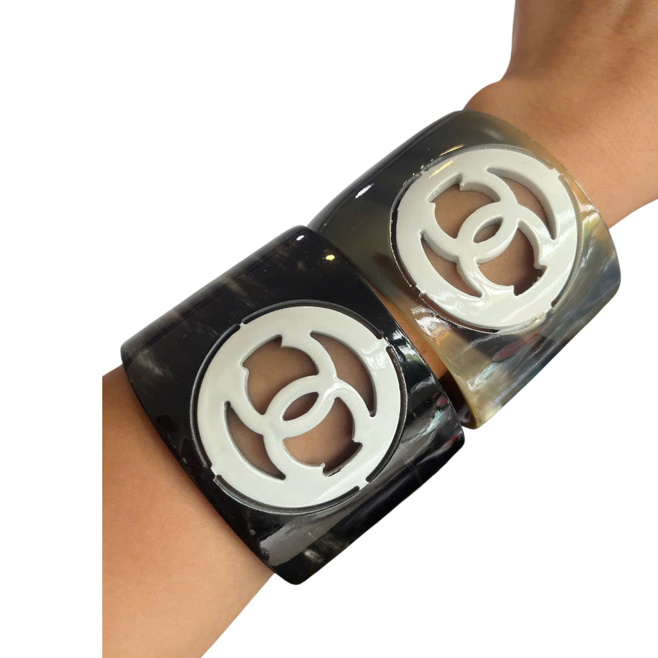 A pair of wide cuff bracelets with a glossy black and smoky gray tortoiseshell pattern, featuring a large white interlocking CC logo cutout in the center, shown worn on a wrist.