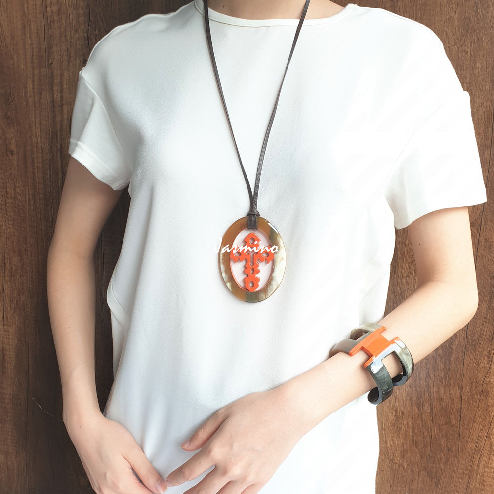 
                  
                    A model is wearing Jasmino unique handmade cross oval pendant in natural buffalo horn and orange for men and women
                  
                